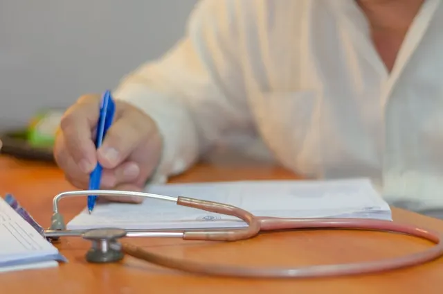 Close-up of a doctor's hands writing notes on a pad with a stethoscope resting on the desk in the foreground.