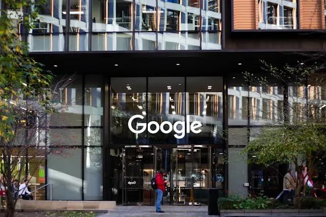 The exterior entrance of a modern Google office building. Large, illuminated white letters spelling "Google" are displayed above glass revolving doors. A person wearing a red jacket and blue jeans stands on the sidewalk in front of the entrance, looking at their phone.