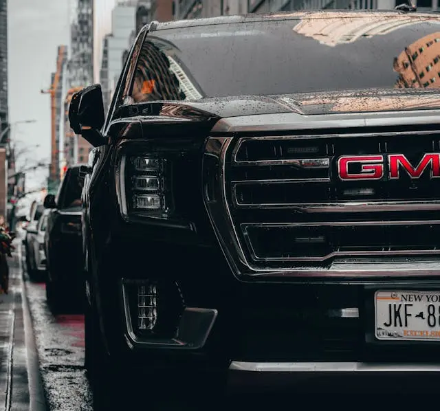 A close-up, low-angle shot of the front of a black GMC truck on a wet city street. The focus is on the large chrome-trimmed grille, the prominent red GMC emblem, and a partial New York license plate, with raindrops scattered across the hood.