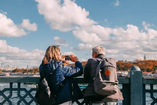 A rear view of a man and a woman leaning on a decorative bridge railing, looking across a wide body of water. The woman is taking a photo with her smartphone, and the man wears a grey backpack. Fluffy white clouds fill the blue sky above a distant, autumn-colored hillside.