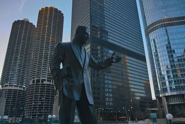 A low-angle shot of a bronze statue depicting a man in a suit gesturing forward. The statue stands in front of a modern cityscape, featuring a tall grid-patterned skyscraper and the distinct cylindrical, corncob-shaped Marina City towers under a clear sky.