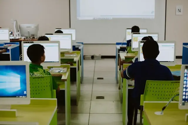 A classroom computer lab with rows of students seated at desks, viewed from behind. The students are working on desktop computers with white monitors and bright lime-green chairs, all facing a large projector screen at the front of the room.